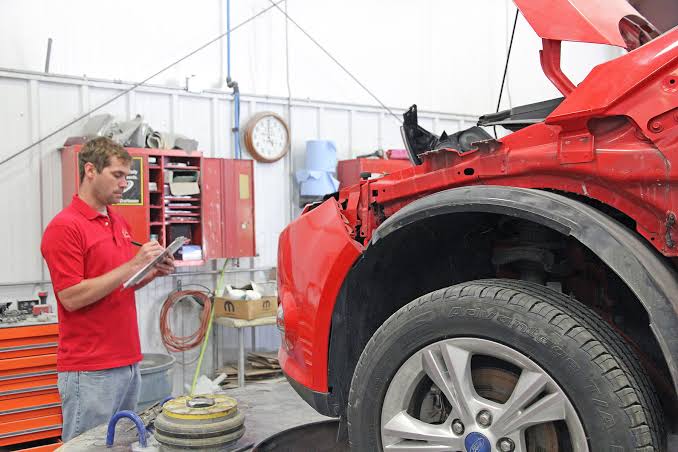 5th & Oak Autobody technician writing a repair estimate beside a damaged red car